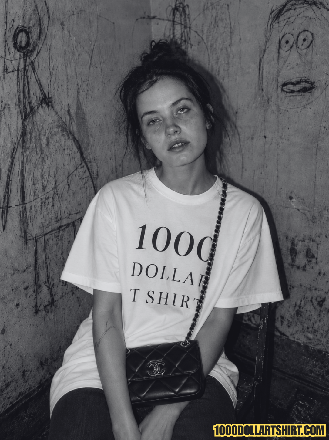 Black and white portrait of a visually impaired woman leaning against a wall with face graffiti, wearing a white t-shirt printed with '1000 DOLLAR T SHIRT' and holding a black quilted handbag.