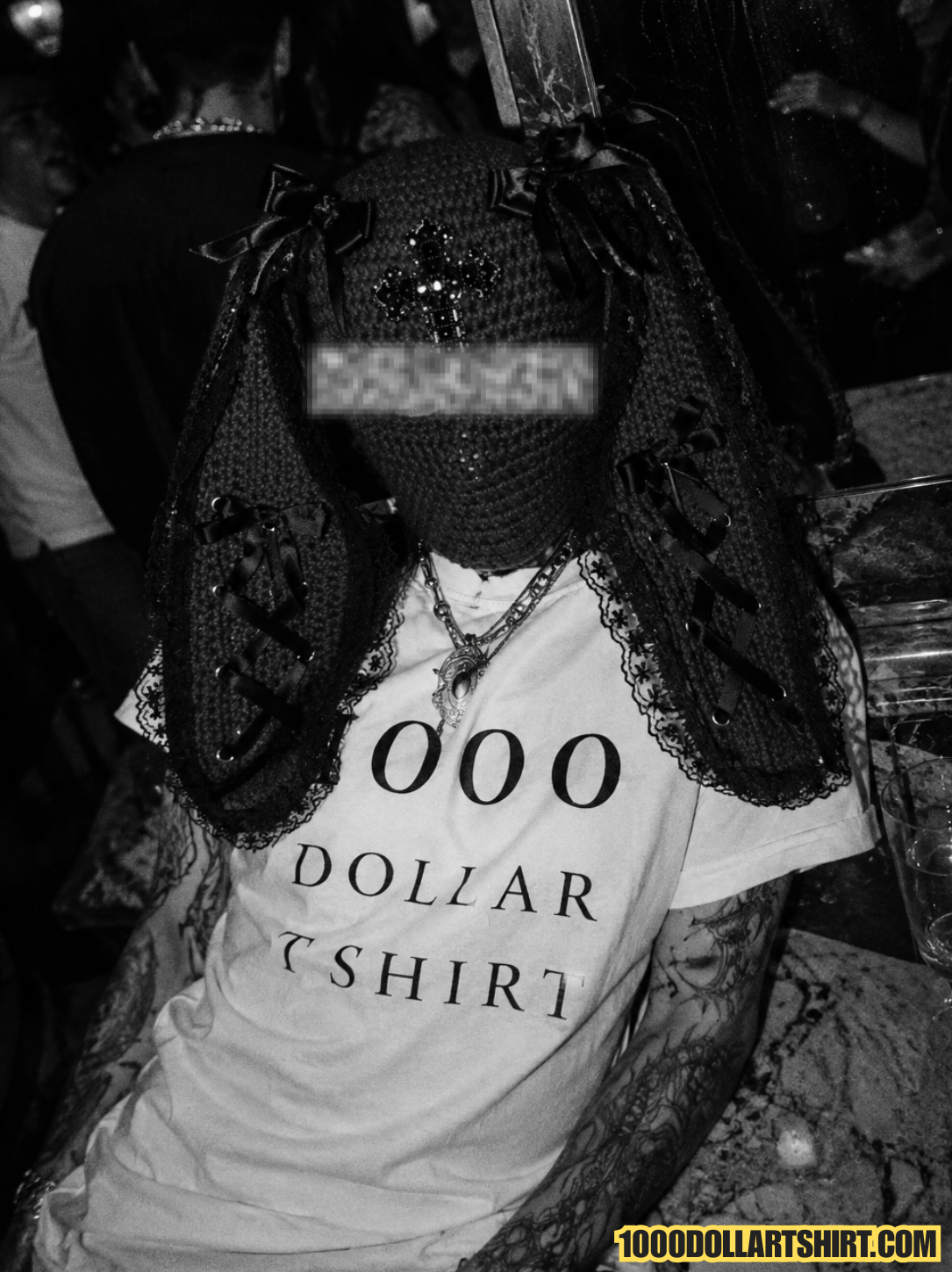 Black and white candid photograph of a woman wearing a 1000 Dollar T Shirt and a gothic bunny balaclava while sitting in a crowded nightclub with layered necklaces and tattooed arms.